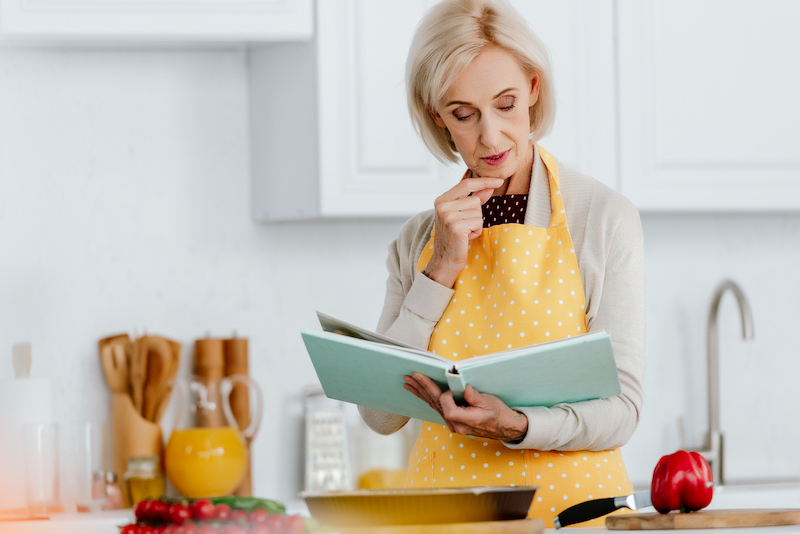 Woman in a yellow polka-dot apron reading a cookbook in her kitchen